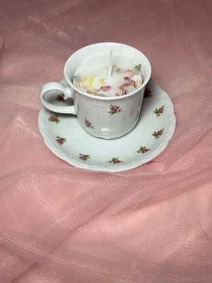 Doftljus i en tekopp i en vit färg med rosa blommor, med torkade blommor pånr 2  - Det här ljuset är gjort av återanvändbara material. Tekoppen är köpt secondhand och stearinet är från kyrkor som tänkte slänga det. Det här ljuset har en svag doft utav vanilj, så känsliga även kan använda det. Det finns även fler ljus med denna design på hemsidan, om man vill köpa sets. Det här ljuset har även torkade blommor på. 