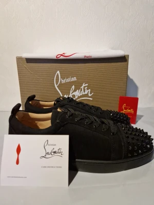 Svarta sneakers från Christian Louboutin - Säljer ett par svarta sneakers från Christian Louboutin med coola svarta nitar på tån och broderad logga på hälen. Skorna har snörning och är tillverkade i mocka med platt sula. Perfekt för dig som vill sticka ut med en lyxig och edgy stil.