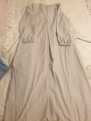 Grå  långklänning med dragkedja - Säljer en stilren grå långklänning i storlek S. Klänningen har rund halsringning, långärmade ärmar med resår vid mudden och en diskret dragkedja framtill. Materialet känns lätt och mjukt, perfekt för dig som gillar enkel och clean stil.