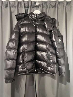 Svart pufferjacka från Moncler - Säljer en svart pufferjacka från Moncler med glansig finish och dunfyllning. Jackan har huva, dragkedja framtill, två fickor med dragkedja och en extra ficka med Moncler-logga på ärmen. Perfekt för kalla dagar och riktigt snygg streetstil. Jackan är äkta dessutom för dem som undrar. Storlek 4. 