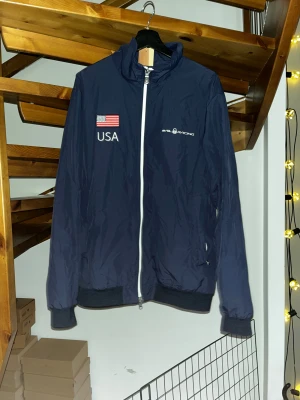 Blå Sail Racing vindjacka USA - Blå vindjacka från Sail Racing med vita detaljer och dragkedja. USA-flagga och text på bröstet samt ryggen. Pris diskuterbart 