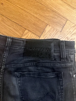 Jacob Cohén Bard Jeans, stl 33 - Snygga svarta jeans från Jacob Cohen med klassisk femficksdesign och svarta detaljer. Jeansen har rak passform och är tillverkade i mjukt jeansmaterial med subtila slitningar framtill. Perfekta för dig som gillar stilrena och tidlösa plagg.