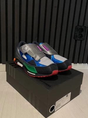 Adidas Raf Simons RS OZWEEGO sneakers, HELT NYA!  - Futuristiska Adidas Raf Simons RS OZWEEGO sneakers i blått, grönt, silver och svart med inslag av lila och orange. Skorna har breda kardborreband, platt sula och coola paneler i syntet och mesh. Perfekt statement-piece för dig som gillar unika detaljer och färg. Finns endast 14 av dessa skor dessa är 5/14! Helt nya skor oanvända!