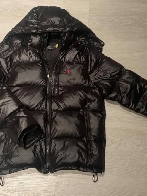 Svart pufferjacka från Polo Ralph Lauren - Svart pufferjacka från Polo Ralph Lauren med röd broderad logga på bröstet. Jackan har huva, två sidofickor med dragkedja och en kraftig frontdragkedja. Materialet är glansigt och känns riktigt mjukt, perfekt för kalla dagar.