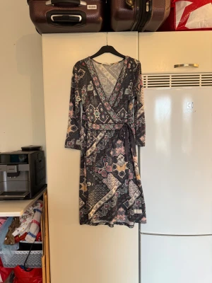 Mönstrad omlottklänning med bälte - Snygg omlottklänning med lång ärm och v-ringning, dekorerad med ett färgstarkt mönster i svart, beige, rosa och turkos. Klänningen har ett knytband i midjan och är tillverkad i ett mjukt, lätt material som ger en skön känsla. Perfekt för dig som gillar boho-vibes och detaljer. Från märket Odd Molly. Storlek 2 som enligt deras storleksguide motsvarar en M. Mycket fint skick, ända anmärkning är att banden har tråddraning. 