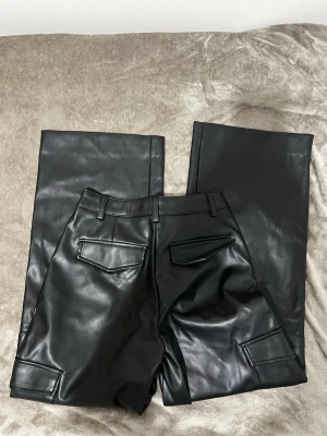 Svarta cargopants i skinnimitation XS - Snygga svarta cargopants i skinnimitation med raka ben och coola fickdetaljer både fram och bak. Byxorna har bälteshällor, dragkedja och en loose passform som ger en trendig vibe. Perfekta för dig som vill sticka ut med en edgy look.
