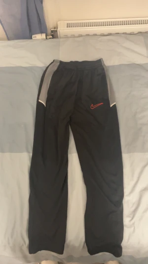 Svarta träningsbyxor från Nike - Svarta träningsbyxor från Nike med grå paneler på sidorna och röd broderad swoosh-logga framtill. Byxorna har elastisk midja och är gjorda i lätt polyester som andas. Perfekta för sport eller chill. Snygg och enkel design med diskreta detaljer.