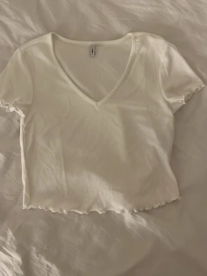 Vit croppad v-ringad topp från Only - Säljer en vit croppad topp från Only med v-ringad hals och korta ärmar. Toppen har vågiga kanter vid ärmslut och nederkant, vilket ger en söt detalj. Perfekt att matcha med jeans eller kjol för en fräsch look.