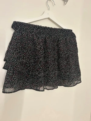 Svart kortkjol med volang från H&M - Säljer en svart kortkjol från H&M med små prickar i vitt och rött. Kjolen har volang och ett smockat midjeband som gör den bekväm och enkel att bära. Materialet är lätt och luftigt, perfekt för varmare dagar. Passar dig som gillar en lekfull och trendig stil. Utstrechad på bilden💕