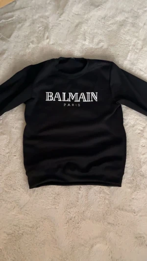 Balmain hoodie Storlek S - Svart sweatshirt från Balmain Paris med vit logotyptryck på bröstet. Tröjan har rund halsringning och långa ärmar. Materialet är mjukt och har en lätt glansig finish, vilket ger en exklusiv känsla. Perfekt för dig som gillar stilrena designer och streetwear.
