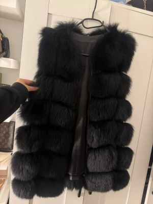 Svart fluffig pälsväst - Säljer en svart pälsväst med riktigt fluffiga och breda sektioner. Västen har en rak och lite oversized passform, och är tillverkad i pälsmaterial som ger en lyxig och cool vibe. Perfekt att slänga över en hoodie eller långärmad tröja för extra värme.