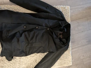 Svart rock från BOSS med knappar - Svart rock från BOSS med klassisk krage och knäppning framtill. Jackan har raka linjer, diskreta fickor och quiltat foder på insidan för extra komfort. Perfekt för dig som gillar stilrena och tidlösa plagg med snygg passform.