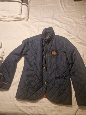 Mörkblå quiltad jacka Morris XXL - Quiltad mörkblå jacka från Morris Lady i storlek XXL. Jackan har guldfärgade knappar, dragkedja, broderad emblem på bröstet och två fickor fram. Materialet är polyester och jackan har en klassisk, lite längre passform med hög krage. Har lite slitage på vissa ställen ( kolla bild) men inget som stör funktionen. Därav nedsatt pris.