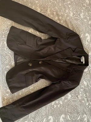 Svart kavaj från Zara Basic - Snygg svart kavaj från Zara Basic med klassisk krage och två knappar framtill. Jackan har två fickor med lock och är helfodrad på insidan. Perfekt för dig som vill ha en stilren och enkel look. Passar till både jeans och kostymbyxor.