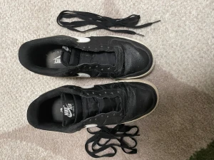 Nike Air Force 1 svartvita sneakers - Svartvita Nike Air Force 1 sneakers med klassisk låg siluett och vit sula. Skorna har svart ovandel i skinn med vita detaljer och logga på hälen. Medföljer svarta skosnören. Perfekt för dig som gillar streetstyle och vill ha en ikonisk sneaker i garderoben.