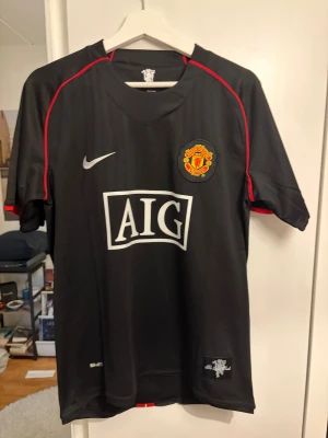 Svarta Nike Manchester United Ronaldo 7 tröja - Säljer en svart Manchester United tröja från Nike med röda detaljer och AIG-logga på bröstet. På ryggen står det Ronaldo och nummer 7 i vitt. Materialet är polyester och tröjan har klubbmärket broderat på framsidan. Perfekt för fotbollsfans!