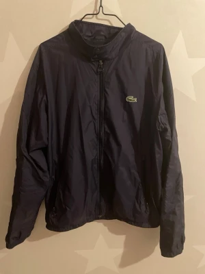 Mörkblå vindjacka från Lacoste - Snygg mörkblå vindjacka från Lacoste med klassisk krokodillogga på bröstet. Jackan har hög krage, hel dragkedja framtill och två sidofickor med dragkedja. Perfekt för dig som gillar stilren och sportig look.