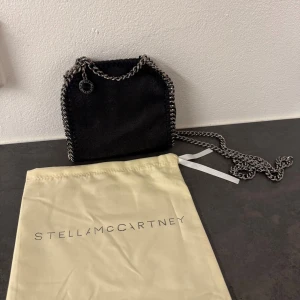 Svart axelväska från Stella McCartney - Säljer en svart axelväska från Stella McCartney med kedjedetaljer i silver runt kanterna och som axelrem. Väskan är i syntetmaterial med en mjuk mockaliknande yta och har en rund logotypbricka i metall. Perfekt för dig som gillar stilrena accessoarer med edge.