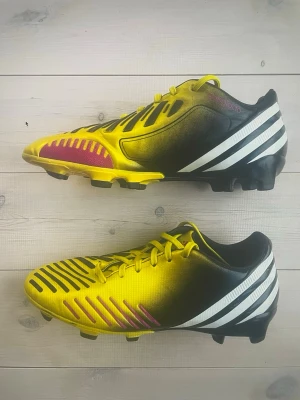 Adidas Predator gula fotbollsskor - Säljer ett par Adidas Predator fotbollsskor i en riktigt fet färgkombo av gult, svart och vitt med rosa detaljer. Skorna har klassiska tre ränder på sidan, snörning och mönstrad ovandel för extra grepp på planen. Perfekta för dig som vill sticka ut på matchen!