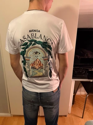 Vit Casablanca Avenida t-shirt - Säljer en vit t-shirt från Casablanca med Avenida-tryck fram och stort grafiskt motiv på ryggen. T-shirten har rund hals och korta ärmar, tillverkad i mjuk bomull. Perfekt för dig som gillar coola prints och streetwear-vibe.