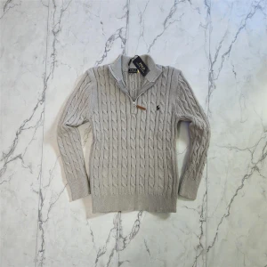 Ljusgrå stickad tröja Polo Ralph Lauren - Snygg ljusgrå stickad tröja från Polo Ralph Lauren med klassiskt kabelstickat mönster och half zip-dragkedja vid kragen. 