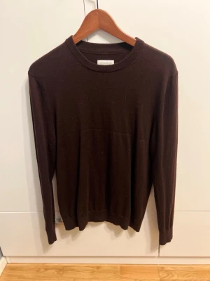 Mörkbrun merinotröja These Glory Days - Snygg mörkbrun stickad tröja från These Glory Days i mjuk superior merinoull. Klassisk rund hals och ribbade muddar vid ärmslut och nederkant. Perfekt för dig som gillar stilrena och enkla plagg med skön passform.