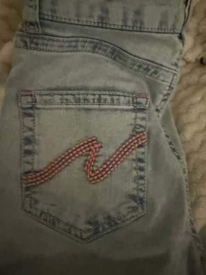 Ljusa ripped jeans från Nelly, strl 36 - Säljer ett par ljusblå jeans från Nelly i storlek 36. De har slitna detaljer och hål på benen, klassisk femficksdesign och rosa broderi på bakfickan. Jeansen har dragkedja och knapp framtill, och är gjorda i jeansmaterial.