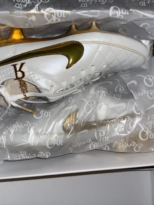 Nike Ronaldinho 10R fotbollsskor vit/guld - Exklusiva Nike Ronaldinho 10R fotbollsskor i vitt skinn med guldfärgade detaljer och sula. Snyggt quiltad ovandel, guldfärgad Nike-swoosh och Ronaldinho 10R-logga. Skorna har snörning och platt sula med dobbar för gräsplan. Perfekt för dig som vill sticka ut på planen.