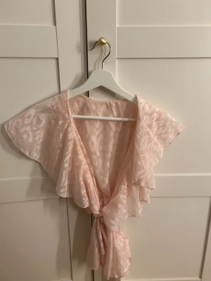 Ljusrosa blus med volangärm - Säljer en ljusrosa blus med korta volangärmar och knytning framtill. Blusen har ett subtilt mönster med glansiga detaljer och är tillverkad i ett lätt, transparent tyg som ger en luftig känsla. Perfekt för dig som gillar romantisk och feminin stil.