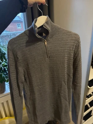 Grå stickad half zip-tröja från Solid - Snygg grå stickad tröja från Solid med half zip-dragkedja i halsen. Tröjan har ett strukturerat mönster och lång ärm, perfekt för lager-på-lager. Hög krage som ger extra stilpoäng och värme under kyliga dagar.