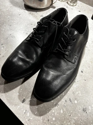 Svarta kostym - skinnskor från Zeus - Stilrena svarta skinnskor från Zeus med klassisk snörning och rund tå. Skorna har en låg klack och är perfekta för dig som vill ha en clean och tidlös look. Tillverkade i slätt skinn som ger en snygg finish.