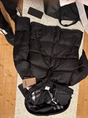 Svart pufferjacka från Burberry - Svart pufferjacka från Burberry med klassisk rutigt foder i grått och vitt. Jackan har dragkedja och knappar framtill, samt flera fickor med dragkedja. Materialet är polyester och jackan har en oversized passform som ger en cool vibe.