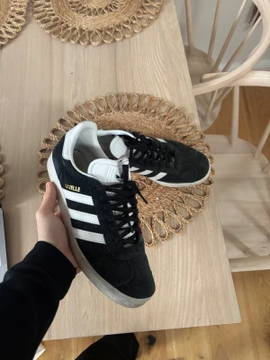 Adidas Gazelle svarta sneakers - Svarta Adidas Gazelle sneakers med vita ränder och vit plös. Skorna har snörning och är tillverkade i mocka med gummisula. Klassisk design med rund tå och låg profil, perfekt för dig som gillar retrostil och streetwear. Storlek 43 1/3 fits 42-43