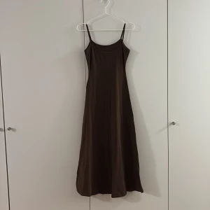 Brun långklänning med smala axelband - Säljer en stilren brun långklänning med smala axelband och rundad halsringning. Klänningen har en enkel och elegant siluett som faller snyggt längs kroppen.