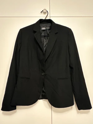 Svart kavaj från Zara, storlek 38 - Klassisk svart kavaj från Zara med enkelknäppning och två fickor framtill. Snyggt skräddad passform och helfodrad insida. Tillverkad i ett mjukt material som känns bekvämt hela dagen.