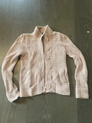 Ljusrosa stickad kofta med stora knappar - Säljer en ljusrosa stickad kofta med hög krage och stora matchande knappar framtill. Koftan har två fickor, ribbade muddar och en snygg, figurnära passform. Perfekt för lager-på-lager och chill höst- eller vinterstil. Passar även S