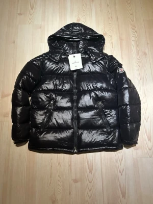 Svart pufferjacka från Moncler- Storlek: M - Svart pufferjacka från Moncler med glansig finish, huva och två snedställda fickor med dragkedja. Klassisk Moncler-logga på ärmen och quiltad design. Jackan är tillverkad i polyester och dun, perfekt för kalla dagar.