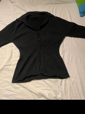 Svart ribbad kofta med krage - Säljer en svart, ribbad kofta med klassisk krage och knappar hela vägen framtill. Koftan är figurnära och har långa ärmar, perfekt för lager-på-lager. Materialet är stickat och känns mjukt mot huden. Passar dig som gillar enkel och stilren look.