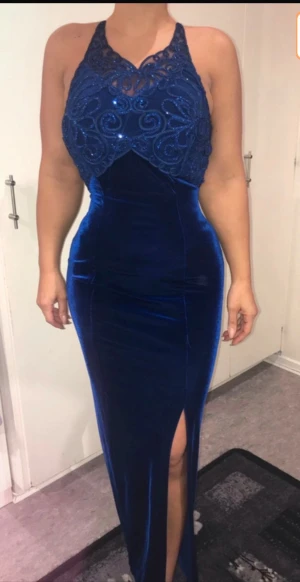 Marinblå långklänning med spets - Säljer en marinblå långklänning i sammet med spetsdetaljer och glittrig brodering upptill. Klänningen har en snygg slits framtill, öppen rygg med knyt och figurnära passform. Perfekt för dig som vill sticka ut med en elegant look.