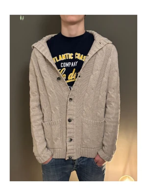 Beige stickad kofta med huva - Säljer en beige stickad kofta med huva och grovt kabelstickat mönster. Koftan har knappar framtill, två stora fickor och förstärkta armbågar i grått tyg. Perfekt för lager-på-lager under kyliga dagar.