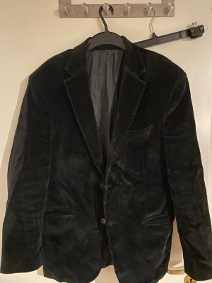 Svart kavaj från Benelli, storlek 56 - Svart kavaj från Benelli i mjuk sammet med klassisk krage och två knappar framtill. Fodrad insida och fickor på sidorna. Perfekt för dig som vill ha en stilren och elegant look. Materialet är 100% bomull och kavajen har en snygg, normal passform.