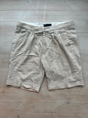 Beige linneshorts från ASOS DESIGN W34 - Snygga beige shorts från ASOS DESIGN i storlek W34. De är tillverkade i ett strukturerat linnematerial med klassisk passform och har både knapp och dragkedja framtill. Perfekta för varma dagar och har en stilren look med diskreta veck fram.