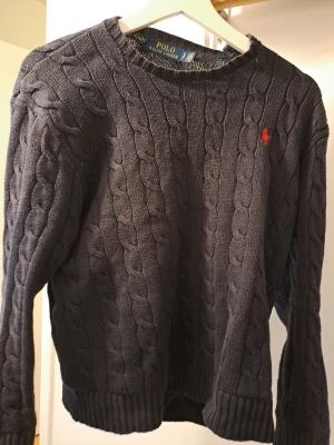 Mörkgrå kabelstickad tröja Polo Ralph Lauren - Kabelstickad mörkgrå tröja från Polo Ralph Lauren med klassisk rund hals och röd broderad logga på bröstet. Tröjan är långärmad och har ribbade muddar vid ärmslut och nederkant. Perfekt för lager-på-lager under kyliga dagar. Skriv prisförslag 