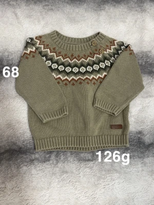Stickad tröja med mönster, storlek 68 - Supersöt stickad tröja från Newbie i grön med klassiskt mönster i vitt, brunt och grönt över bröstet. Tröjan har rund halsringning, två träknappar på axeln och långa ärmar. Perfekt för kyliga dagar!🌸 Fint använt skick. Storlek 68