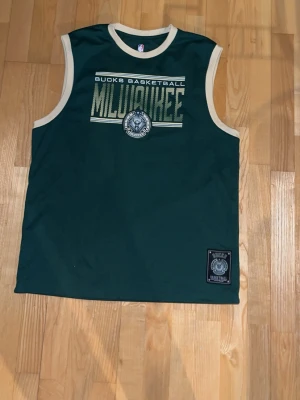 Milleuakee bucks träningslinne  - Grönt ärmlöst linne från Milwaukee Bucks med vit kantning. Tryck med “Bucks Basketball Milwaukee” och lagets emblem på bröstet samt NBA-detalj nedtill. Sportig och klassisk design.