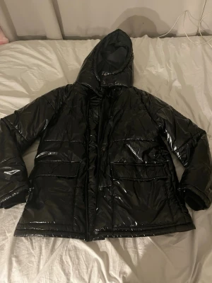 Svart glansig pufferjacka från Boohoo - Mest intressekoll men Säljer en svart, glansig pufferjacka från Boohoo med huva och stora fickor framtill. Jackan har dragkedja och tryckknappar. Jag är 158 cm.  Huvan är reflex inuti.