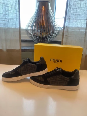 Svarta Fendi sneakers - Säljer ett par svarta Fendi sneakers med ikoniskt FF-monogram i grått på sidan. Skorna har vita platta sulor, svarta skosnören och detaljer i mocka och skinn. Klassisk låg modell med rund tå och snygg kontrast mellan material och färger. Kommer med originalkartong.