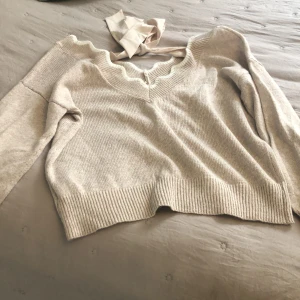 Beige stickad tröja med rosett - Supersöt beige stickad tröja med lång ärm och ribbade muddar. Tröjan har en v-ringad hals med vågig kant och en stor rosett i nacken som ger en extra gullig touch. Perfekt för dig som gillar mjuka och feminina detaljer.