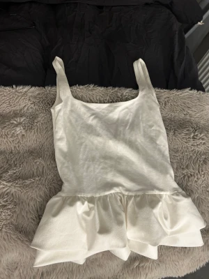 Vit volangtop med breda axelband - Säljer en vit topp med breda axelband och volang nedtill. Toppen har en enkel, stilren design med en lätt glansig finish och är ärmlös. Perfekt för dig som gillar en feminin och trendig look.