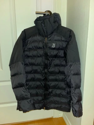 Svart Haglöfs Essens Mimic Parka dam - Säljer en svart Essens Mimic Parka från Haglöfs för dam. Jackan har huva, quiltad design och dragkedja framtill. Perfekt för kalla dagar med sin långa passform och lätta syntetfyllning. Stilsäker och funktionell för vinter och höst. PRIS KAN DISKUTERAS!